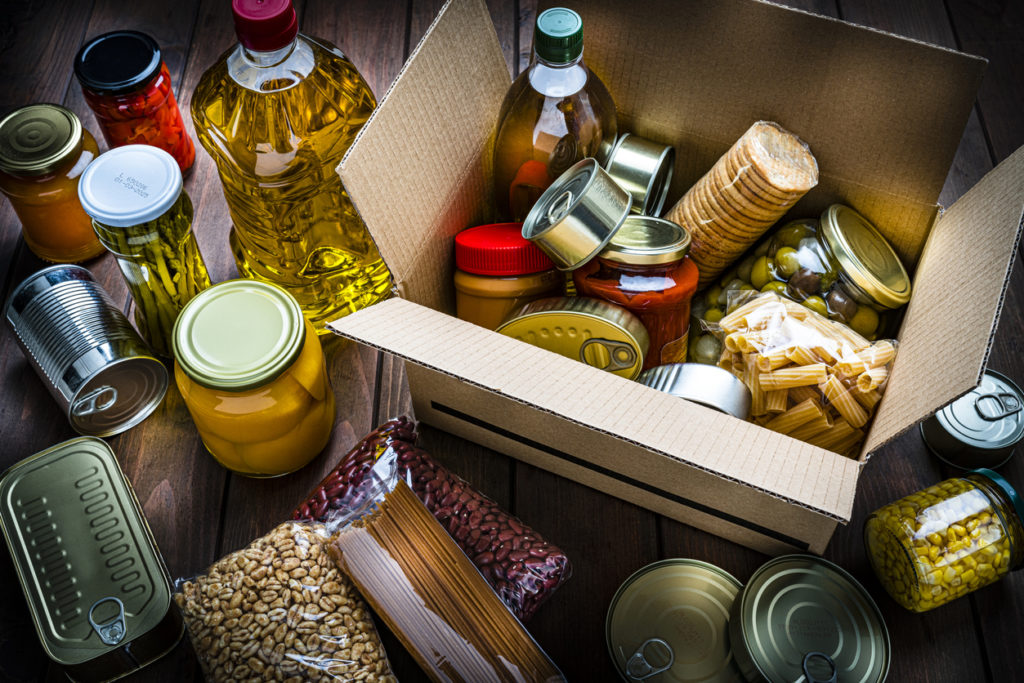High angle view of a cardboard box filled with multicolored non-perishable canned goods, conserves, sauces and oils shot on wooden table. The composition includes cooking oil bottle, pasta, crackers, preserves and tins.