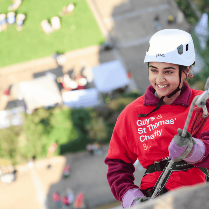 Woman appears to be smiling holding abseil rope, abseiling down St Thomas hospital with the hospital garden in the background below her.
