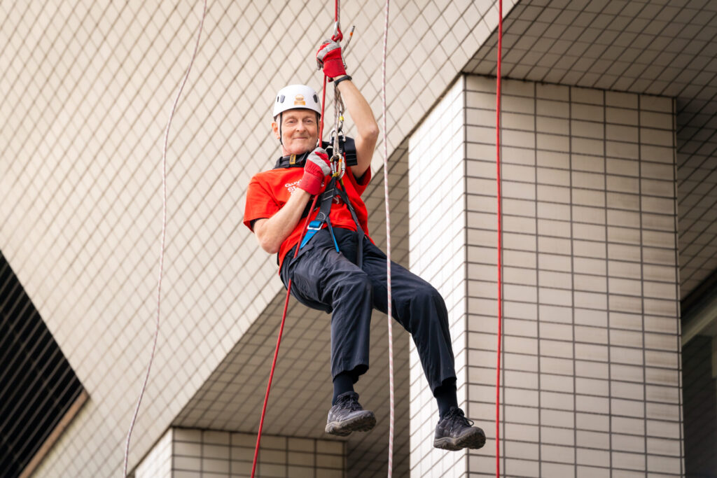 Nigel mid air in abseil harness, holding onto the abseil rope wearing a helmet, in front of St Thomas Hospital.