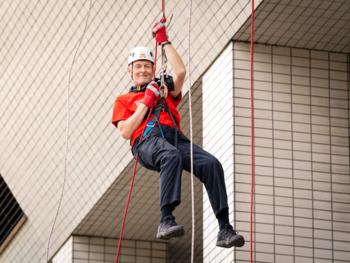 Nigel mid air in abseil harness, holding onto the abseil rope wearing a helmet, in front of St Thomas Hospital.