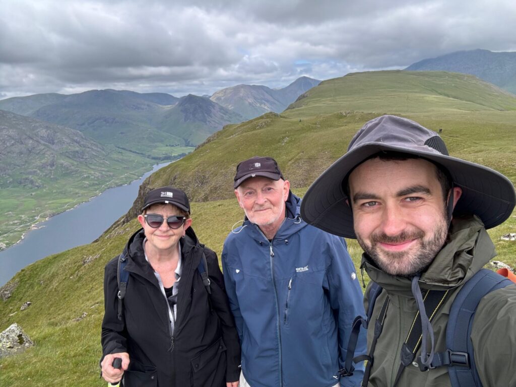 Nigel in the middle with his wife on the left and his son on the right, all are wearing hats with green grass hilltops behind them.