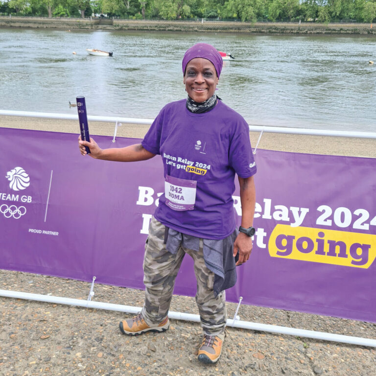 Dioma smiling in a purple headscarf and t shirt holding a purple relay baton in her right hand, in front of a river.