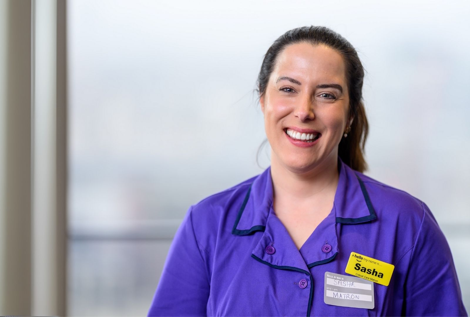 Sasha appears to be smiling at St Thomas' hospital, she's wearing purple uniform with a yellow name badge that reads 'Hello my name is Sasha'.