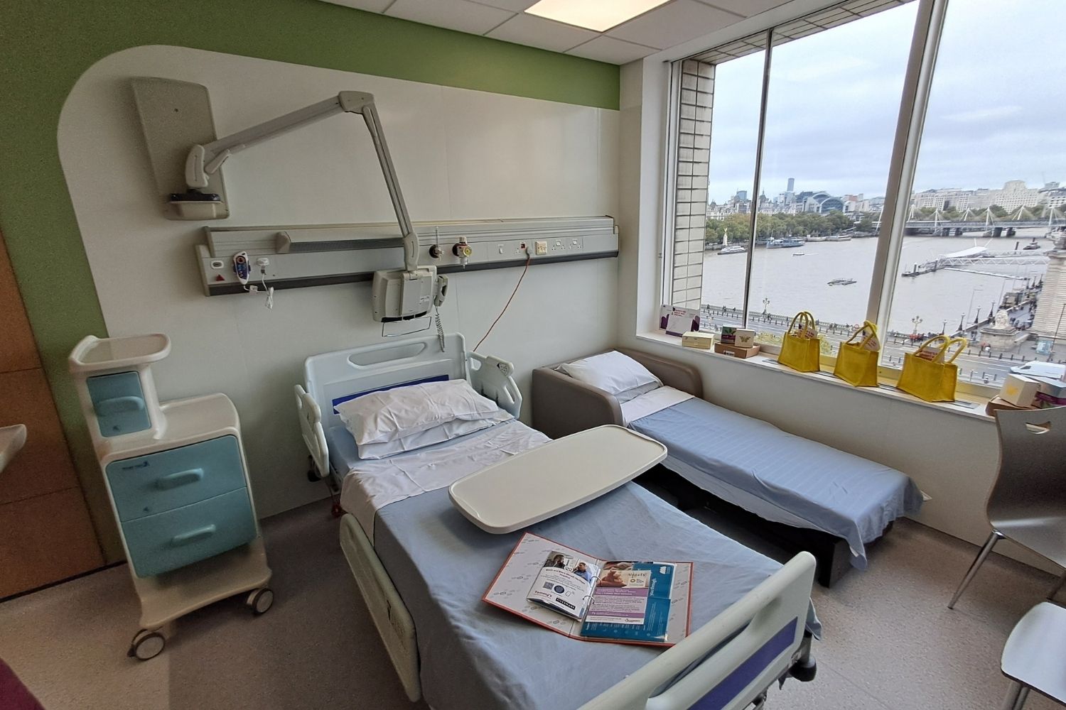 Two beds with blue blankets on the top in the early pregnancy bereavement suite in St Thomas' Hospital Gynaecology unit, on the window sill there are small yellow bags and bereavement resources on the left side.