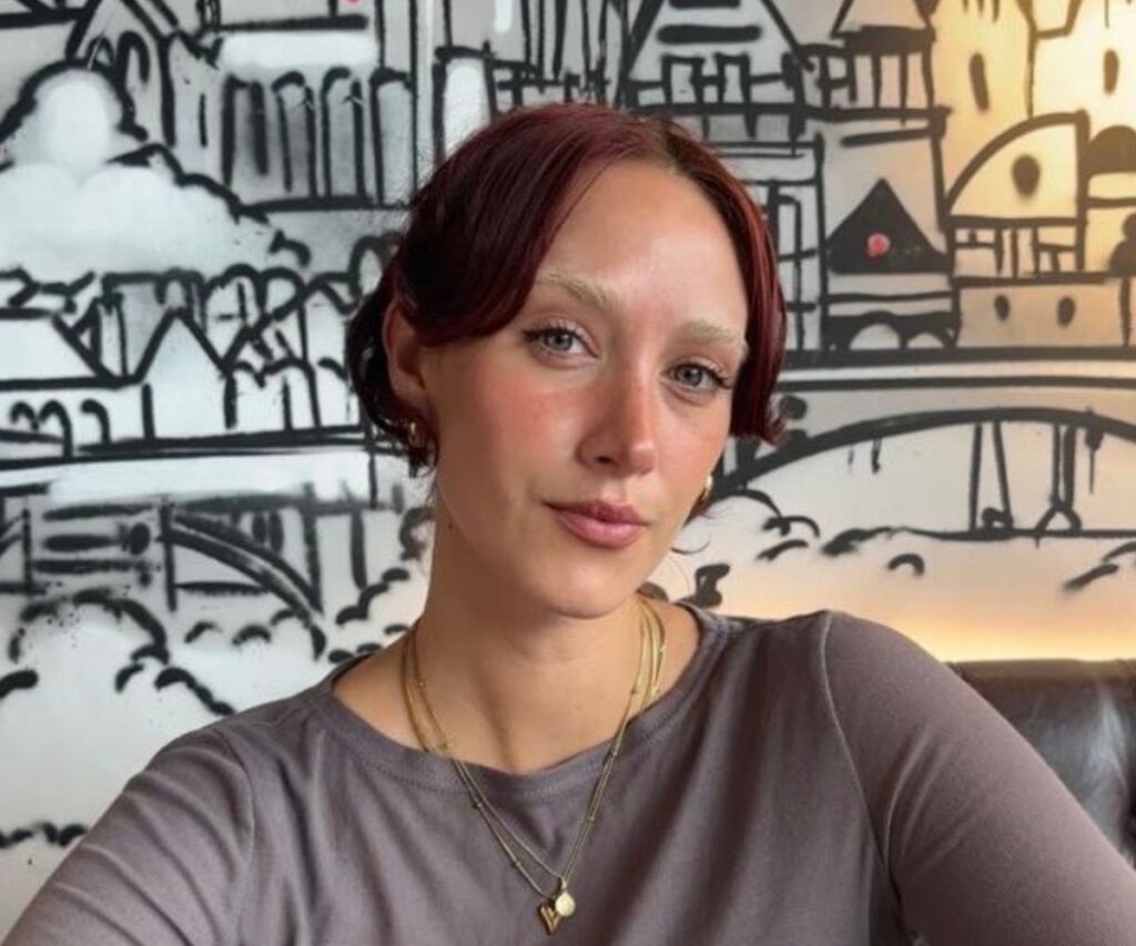 Woman with dark brown hair tied in a bun, wearing a brown top and a gold pendant necklace, sat in front of a white wall featuring a black graphic illustration of buildings.