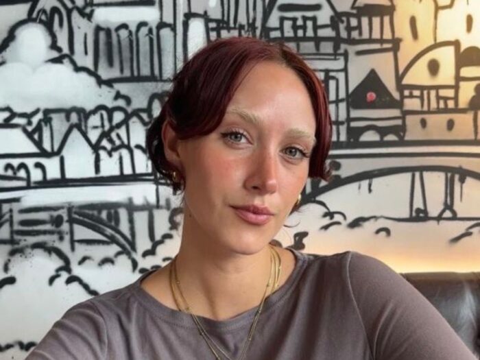 Woman with dark brown hair tied in a bun, wearing a brown top and a gold pendant necklace, sat in front of a white wall featuring a black graphic illustration of buildings.