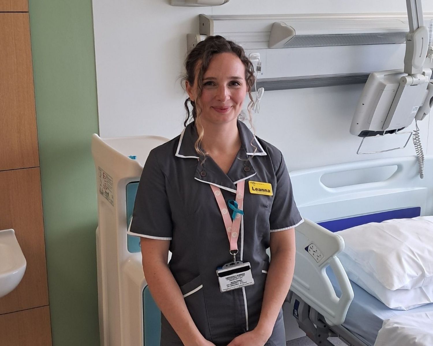 Leanna, a research nurse and early pregnancy specialist at Guy's and St Thomas' hospital, she appears to be smiling and is wearing a grey uniform and wearing a yellow badge that reads 'Leanna' and a light pink lanyard for her staff pass.