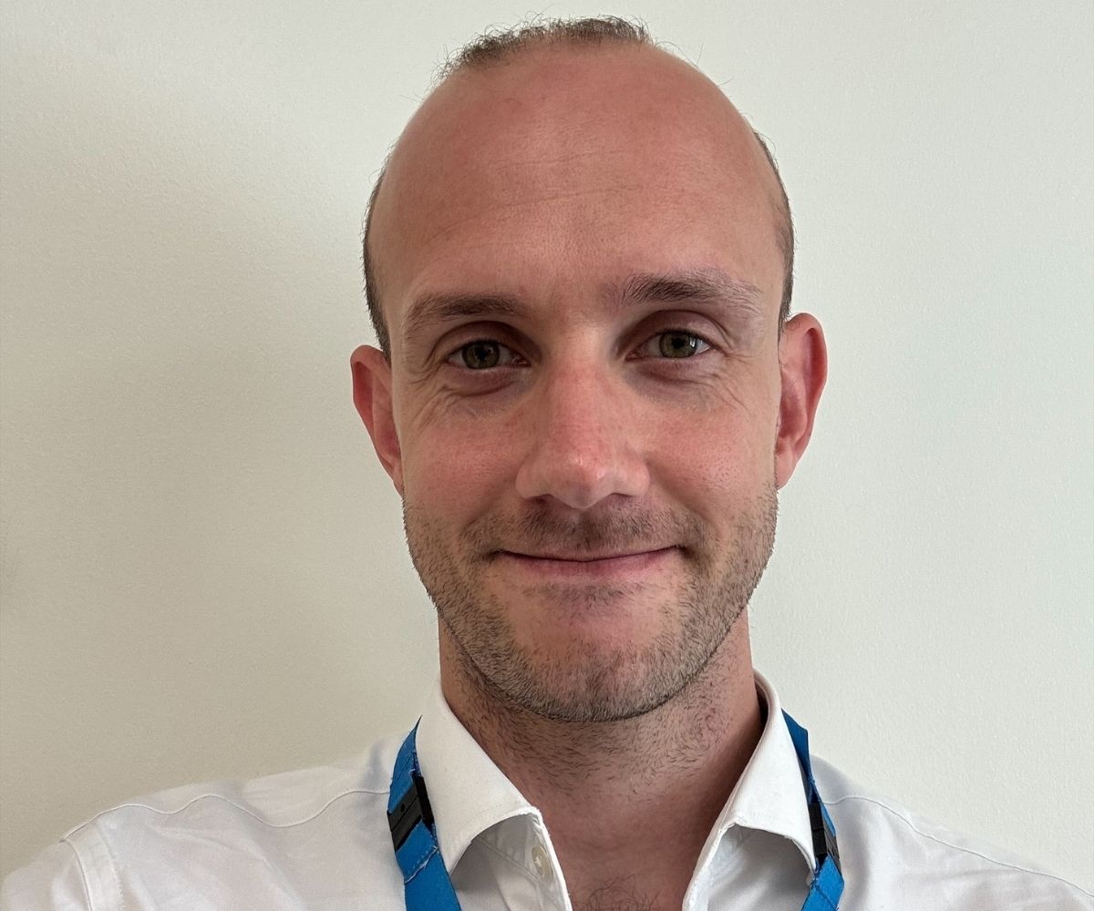 A headshot of George, looking forward smiling in a a white shirt with a blue Guy's and St Thomas' NHS lanyard.