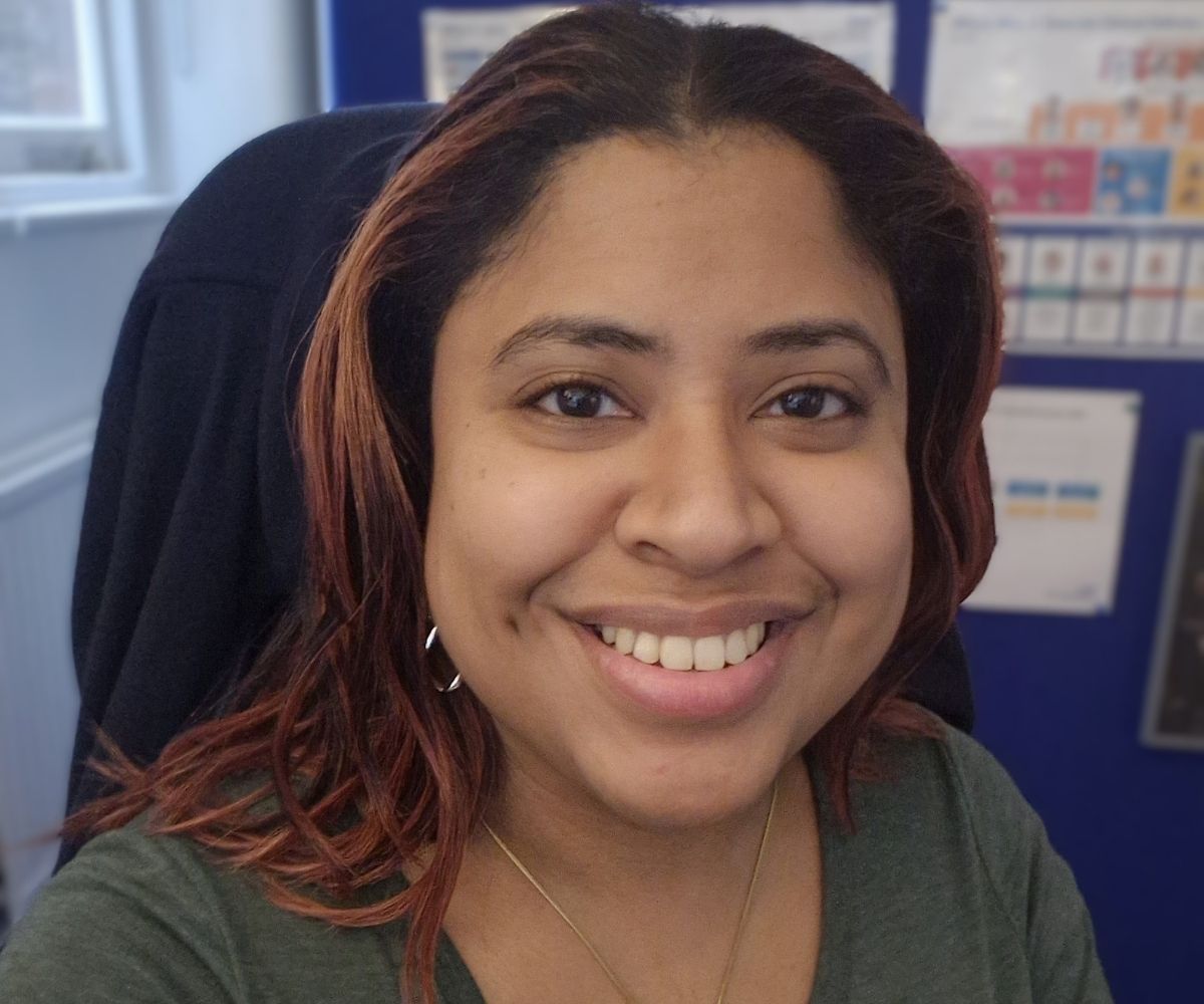 A portrait shot of Sabrina, she appears to be smiling, she has dark wavy hair with highlights around her face, she's in a green jumper and in the background there are infographic posters.