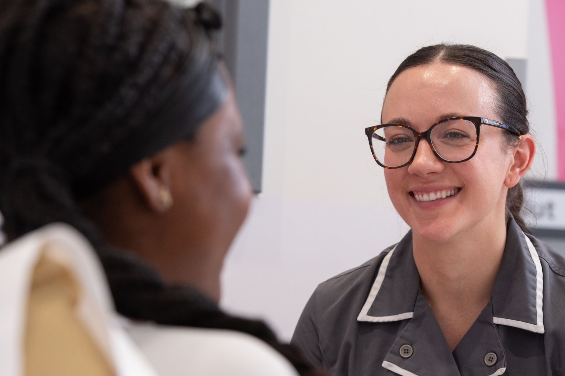 Guy's and St Thomas' nurse, Hannah, in a grey uniform with white piping on the collar, they're wearing glasses with a dark frame and they appear to be smiling at a patient we can see the back of on the left.
